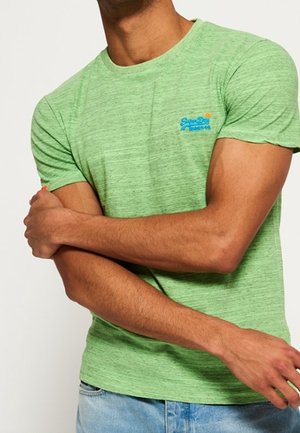 Homme croisant les bras, portant un t-shirt à manches courtes vert chiné avec un petit logo bleu et orange sur le côté gauche de la poitrine, associé à un jean bleu clair.