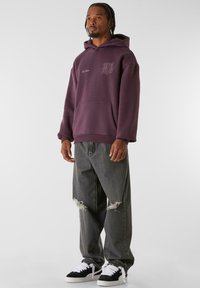 Burgunderfarget hettegenser med frontlomme, med grafisk tekst. Kombinert med grå baggy jeans som har rifter. Svart-hvite sneakers.