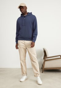 Felpa blu navy con cordini, con un piccolo testo bianco "HUG". Abbinata a pantaloni arrotolati color crema e sneaker bianche.