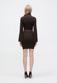 Brun, struktureret kjole med høj krave, lange klokkeærmer og en tætsiddende silhuet. Inkluderer et kædebælte. Modellen har sorte hæle på.