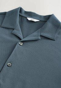 Polo shirt in gedempt teal, met een klassieke kraag, een gestructureerde stof en twee knopen bij de sluiting. Zachte, gladde textuur.