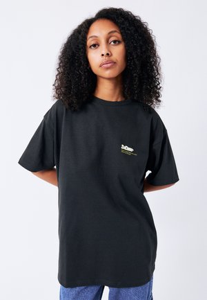 Jonge vrouw met krullend haar die een zwart oversized t-shirt draagt met een klein lichtkleurig logo op de borst en een blauwe spijkerbroek.