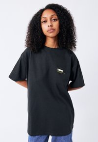 Černé bavlněné tričko s volným střihem, krátkými rukávy a malým zlatým logem na hrudi. Hladká textura, neformální design.