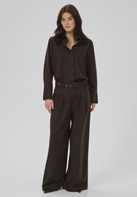Nainen, joka on pukeutunut tummanruskeaan pinstripe-painettuun nappipaitaan, joka on piilotettu yhteensopiviin leveälahkeisiin housuihin ja vyöhön, seisoo vaaleanharmaata taustaa vasten.