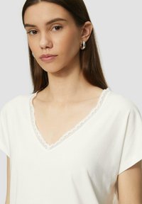 Weißes V-Ausschnitt-T-Shirt mit kurzen Ärmeln, versehen mit zarter Spitzenborte entlang des Ausschnitts. Glattes Baumwollmaterial mit minimaler Textur.