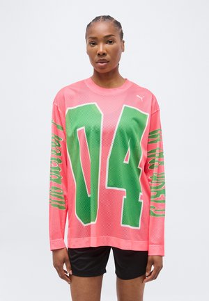 Vrouw draagt een felroze mesh shirt met lange mouwen, met een grote groene "04" op de voorkant en bijpassende groene tekst op de mouwen, gecombineerd met een zwarte korte broek.