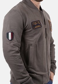 Giubbotto bomber grigio con zip frontale, polsini a coste, due tasche laterali e toppe ricamate tra cui una bandiera italiana e la scritta "LEADER".