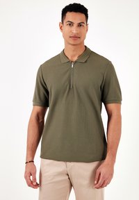 Olivově zelená polokošile se zipovým límečkem, krátkým rukávem a strukturovanou tkaninou. Model má na sobě khaki kalhoty.