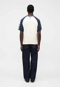 T-shirt με μανίκια ράγκλαν σε εκρού και ναυτικό μπλε, με κόκκινη βούλα στην πλάτη, φορεμένο με σκούρο τζιν παντελόνι που έχει τσέπες τύπου κάργκο.