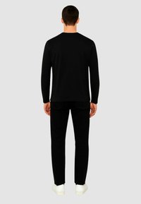 Maglietta nera a maniche lunghe con scollo rotondo, abbinata a pantaloni neri e sneakers bianche, vista da dietro su uno sfondo grigio.