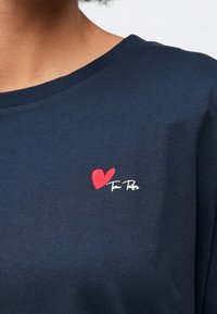 Σκούρο μπλε πουκάμισο με μια μικρή κόκκινη κεντητή καρδιά και λευκό κείμενο "Tom Tailor" στο άνω μέρος του στήθους κοντά στον λαιμό.