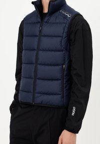 Mężczyzna w granatowej, ocieplanej kamizelce nosi czarną koszulę z długim rękawem z logo "HUGO" na ramieniu i rękawie.