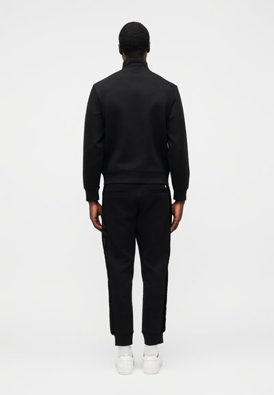 Juodas sportinis kostiumas su aukštu apykakliu; džemperyje yra ribotieji rankogaliai; kelnės turi šonines juostas ir siaurėjančias kojas. Baltos sportbačiai.