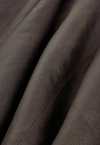 Tessuto in pelle marrone scuro con una fine trama visibile e cuciture a vista, mostrato in primo piano con pieghe morbide.