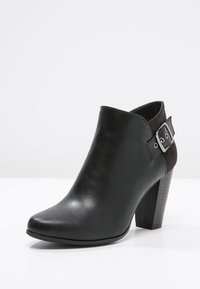 Stivaletto in pelle nera fino alla caviglia con punta affusolata, caratterizzato da un accento in camoscio e dettagli con fibbia argentata sul lato, e tacco impilato.