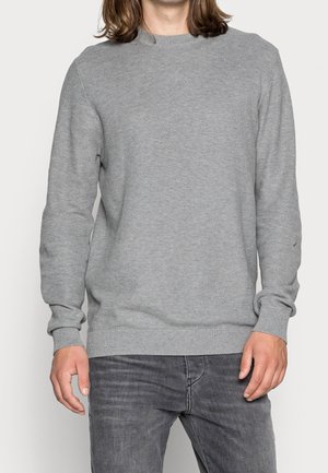 Mann trägt einen hellgrauen, strukturierten Pullover mit langen Ärmeln und dunkelgraue Jeans, steht vor einem schlichten hellen Hintergrund.