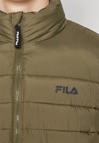 Olivgrön vadderad jacka med hög krage, dragkedjestängning och Fila-logotyp. Har horisontell sömnad och en mjuk textur.
