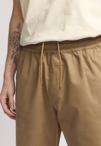 Tan elastische broek met een tailleband met trekkoord, voorzien van zijzakken en een gladde textuur. Versterkt met gouden uiteinden op de trekkoorden.