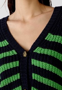 Námořnicky modrý a zelený pruhovaný kardigan s V-výstřihem, žebrovanou texturou a jedním kulatým knoflíkem u výstřihu. Měkká pletená látka.