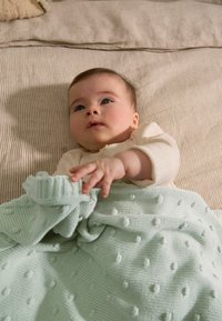 Couverture tricotée vert menthe avec des pois texturés, drapée sur un tissu rayé beige clair. Un bébé en tenue crème interagit avec la couverture.