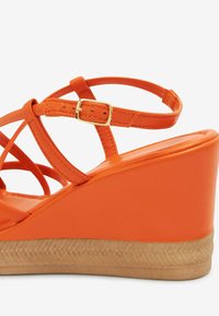 Sandale orange à talon compensé avec des brides croisées, une fermeture à boucle, une tige synthétique lisse et une semelle extérieure texturée pour une meilleure adhérence.