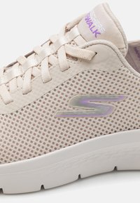 Baskets en maille beige avec un design léger, des lacets fins et une semelle en caoutchouc blanc. Logo notable aux couleurs irisées sur le côté.