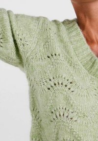 Pull tricoté vert clair avec un col en V, arborant un motif ondulé délicat et une finition douce et texturée.