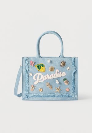 Sac cabas bleu clair à bords à franges, orné de patchs brodés colorés et d'épingles, incluant le mot « "Paradise" » et des motifs tropicaux.