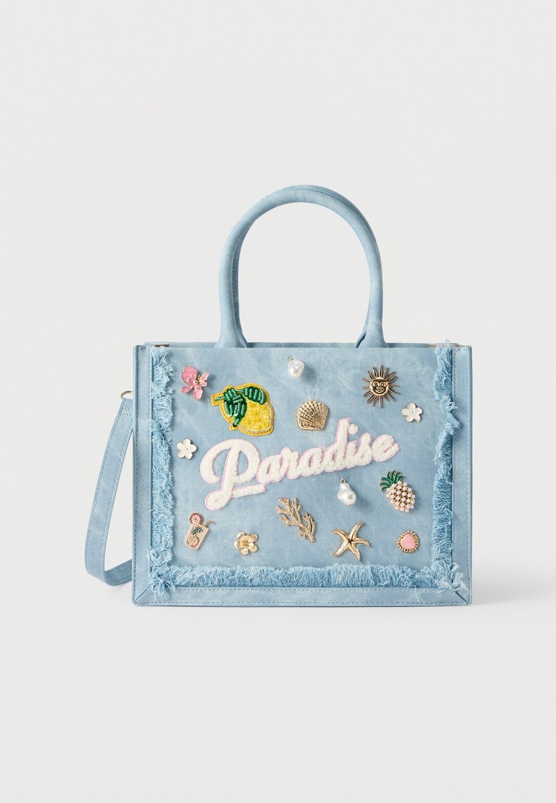 Sac cabas bleu clair à bords à franges, orné de patchs brodés colorés et d'épingles, incluant le mot « "Paradise" » et des motifs tropicaux.
