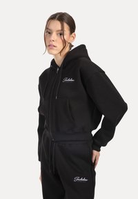 Haut à fermeture éclair noir en tissu doux, doté d'une poche avant et d'une broderie de logo sur la poitrine. Assorti avec un pantalon de jogging noir.
