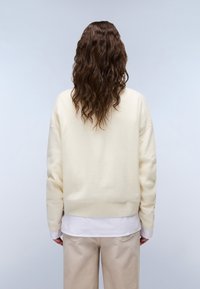 Person med langt, bølget brunt hår iført en cremefarvet sweater, hvid skjorte og beige bukser, der står med ryggen til mod en ensfarvet baggrund.