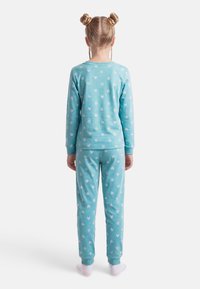 Παιδί που στέκεται με την πλάτη, φοράει ταιριαστά τιρκουάζ πιτζάμες με λευκά σχέδια καρδιών και λευκές κάλτσες σε απλό λευκό φόντο.