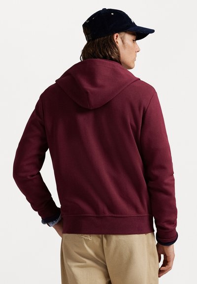 Sweat à capuche bordeaux avec cordon de serrage, texture douce, poignets et ourlet côtelés, porté avec un pantalon beige, avec une casquette de baseball sur la tête.