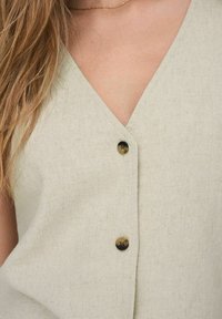 Světlý zelený lněný top s výstřihem do V, zdobený třemi hnědými knoflíky na přední straně a texturovaným látkovým povrchem.