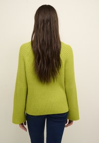 Pull en maille côtelée vert lime avec manches larges, ourlet avant légèrement plus long que l'arrière, associé à un jean bleu foncé, tissu texturé.