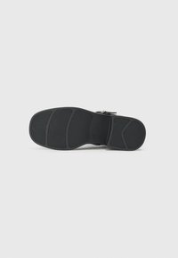 Semelle de sandale noire en caoutchouc texturé pour une meilleure adhérence, dotée d'une forme incurvée et de motifs de bande de roulement distincts. Accent de boucle près du talon.