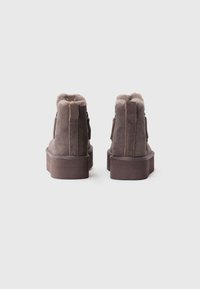 Par de botas de ante marrón hasta el tobillo, con forro de piel sintética y suelas gruesas de plataforma, vistas desde atrás sobre un fondo blanco sencillo.
