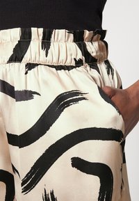 Detailní záběr pasu osoby, která nosí vzorované kalhoty s černými štětci na světlém pozadí, ruka odpočívající v kapse.