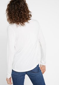 Langärmliges weißes Oberteil mit abgerundetem Saum, aus weichem Stoff mit glatter Textur, von hinten gesehen, kombiniert mit dunkelblauen Jeans.