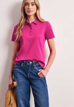 Kobieta w jasnoróżowej koszulce polo i niebieskich dżinsach, trzymająca jasnobrązową torebkę, stoi na tle jasnej ściany.