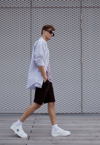 Model nosi niebiesko-białą koszulę w paski na jasnofioletowej bluzce, czarne szorty i białe sneakersy z niebieskimi i czerwonymi akcentami.