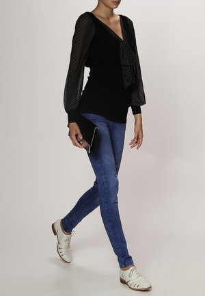 Schwarzes Oberteil mit gepunkteten, durchsichtigen Ärmeln, Schleifendetail, kombiniert mit blauen Slim-Fit-Jeans und weißen Schnürschuhen, hält eine schwarze Clutch.