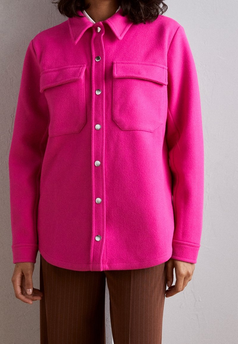 Fleece košile v jasně růžové barvě s předním zapínáním na knoflíky, dvěma kapsami na hrudi a stříbrnými patentkami. Dlouhé rukávy s detaily na manžetách.