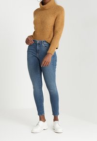 Brun stickad polotröja, kombinerad med blå skinny jeans och vita skor. Kläderna förenar en strukturerad topp med figurnära byxor.