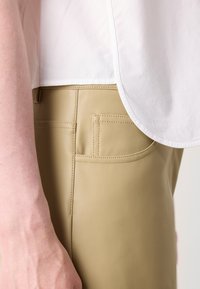 Lyse beige skinnbukser med glatt tekstur, med to baklommer. En hvit topp overlapper midjebåndet, delvis synlig.