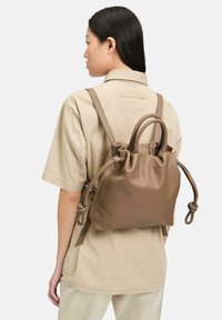 Brązowy skórzany plecak z zaciąganym wierzchem, dwoma szelkami oraz małym, okrągłym uchwytem. Posiada gładką fakturę i węzłowe akcenty.