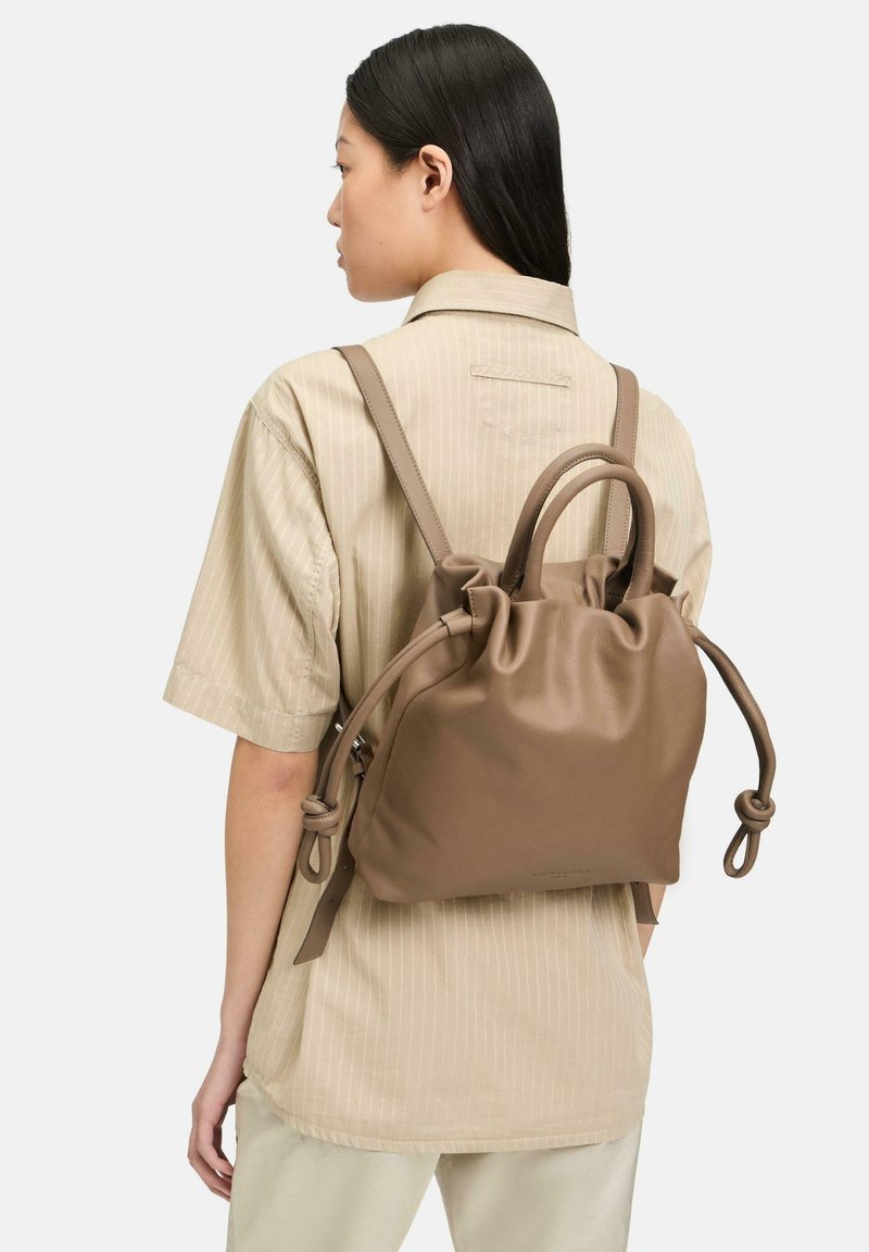 Brązowy skórzany plecak z zaciąganym wierzchem, dwoma szelkami oraz małym, okrągłym uchwytem. Posiada gładką fakturę i węzłowe akcenty.