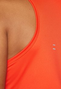 Close-up van de schouder van een persoon die een mouwloos feloranje topje draagt met twee kleine reflecterende rechthoeken vlakbij de naad.