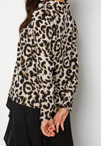 Vrouw met een lange mouwen beige blouse met zwart en bruin luipaardprint, handen achter de rug gekruist, gecombineerd met een zwarte rok.