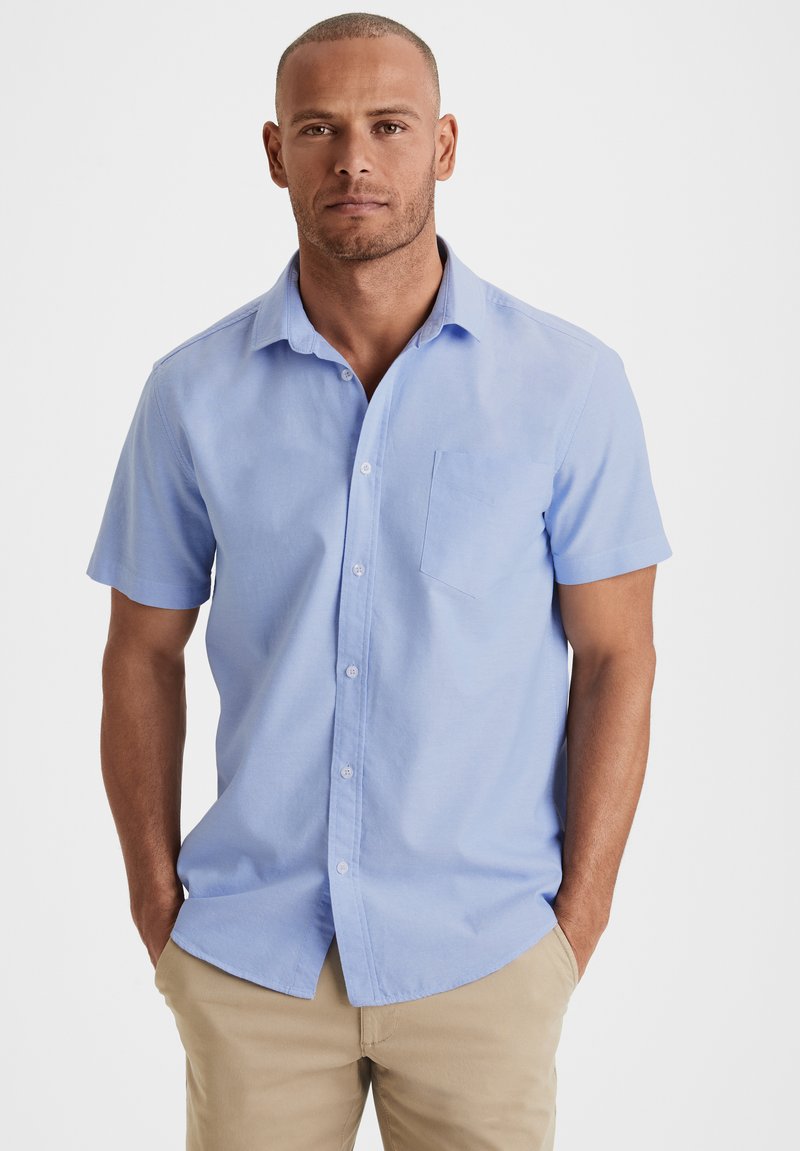 Hellblaues kurzärmeliges Button-Up-Hemd mit einer Brusttasche, aus einem Stoff mit feiner Textur. Das Model posiert mit den Händen in den Taschen.